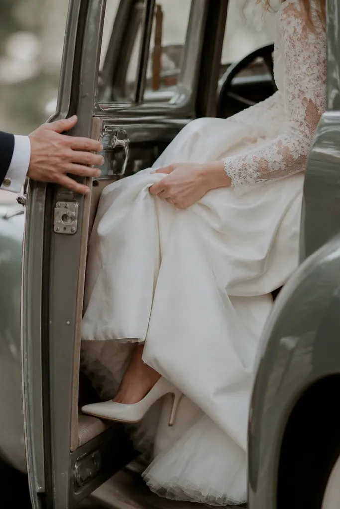 A vintage car with the door opening; the groom’s hand holds the door edge while the bride steps in, with the camera low and focused on hands and fabric details.