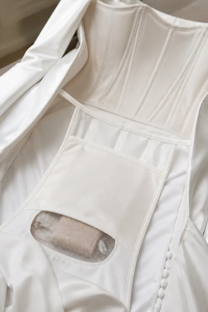 The inside of a long-sleeve wedding dress is shown with a structured bodice and a pocket panel sewn into the inner skirt. A small mesh opening reveals a compact kit compartment.