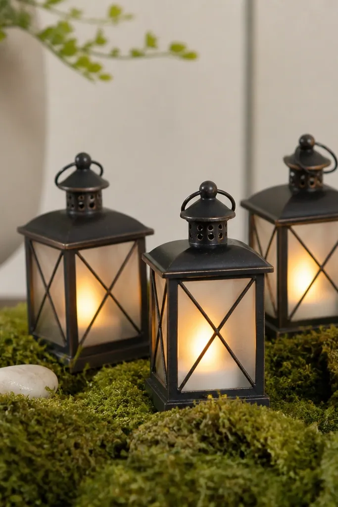 Three small metal lanterns with frosted panels sitting on a bed of faux moss, each glowing with an LED candle, with a couple of white pebbles tucked in.