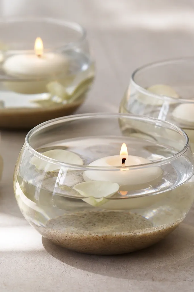 Clear glass bowls filled with water and fine sand, each holding a floating candle, with a few small cream rose petals floating on top.