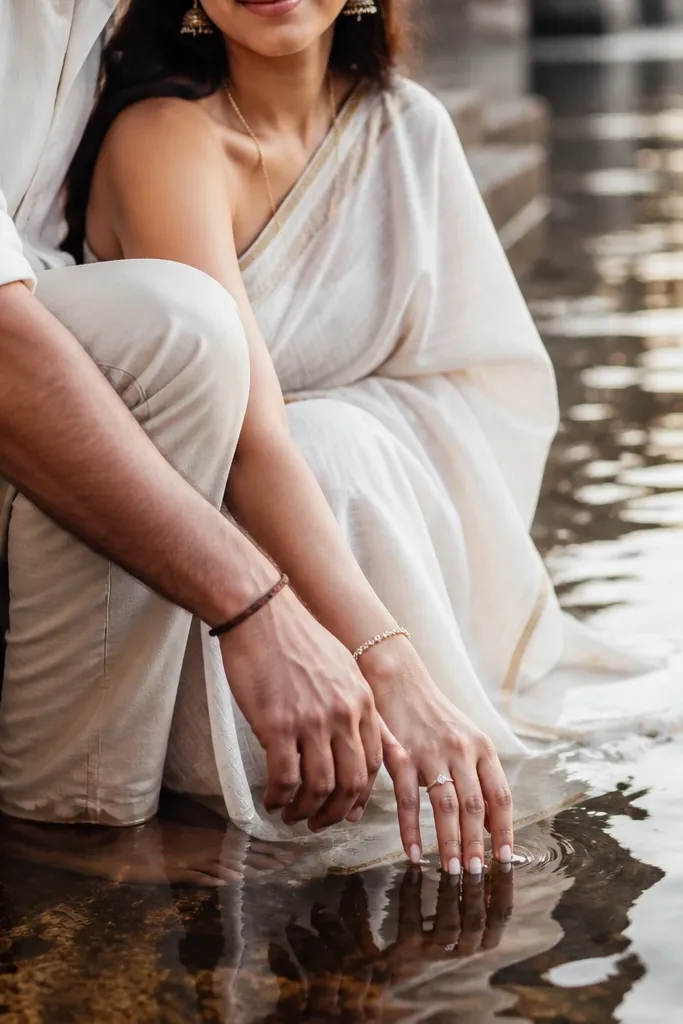 The couple sits on river steps with gentle water reflections; close-up framing shows their hands and jewelry near the waterline; soft ripples blur the background.