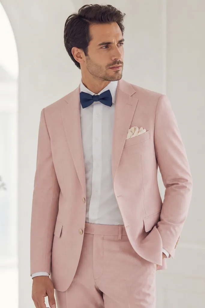 A blush pink suit in a matte fabric, with a matching jacket and trousers. A navy bow tie stands out at the collar, and a pale pocket square peeks from the breast pocket. The background is light and airy.